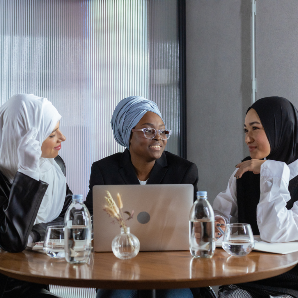 3 women wearing hijabs are sitting at a table looking over a laptop together.